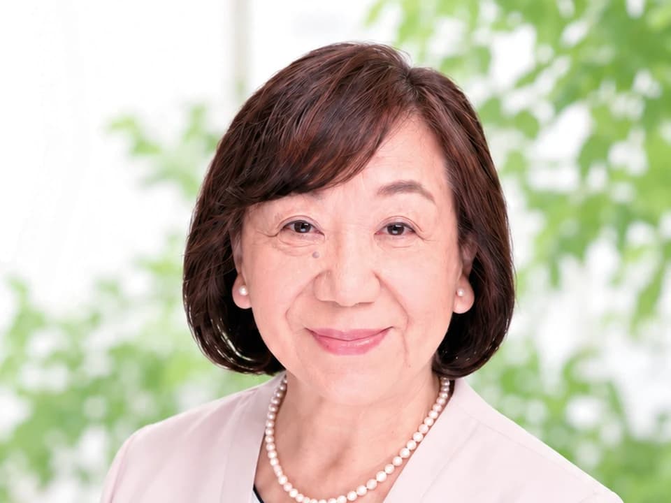清田のり子の写真。やわらかい緑の窓辺で、真珠のネックレスをつけてほほえんでいる